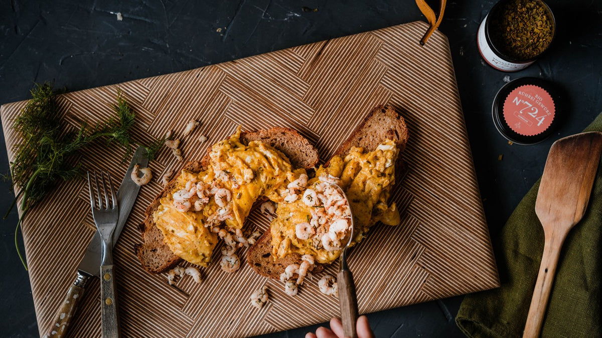 Rührei und Garnelen auf Toast mit Kräutern, serviert auf einem gemusterten Brett.