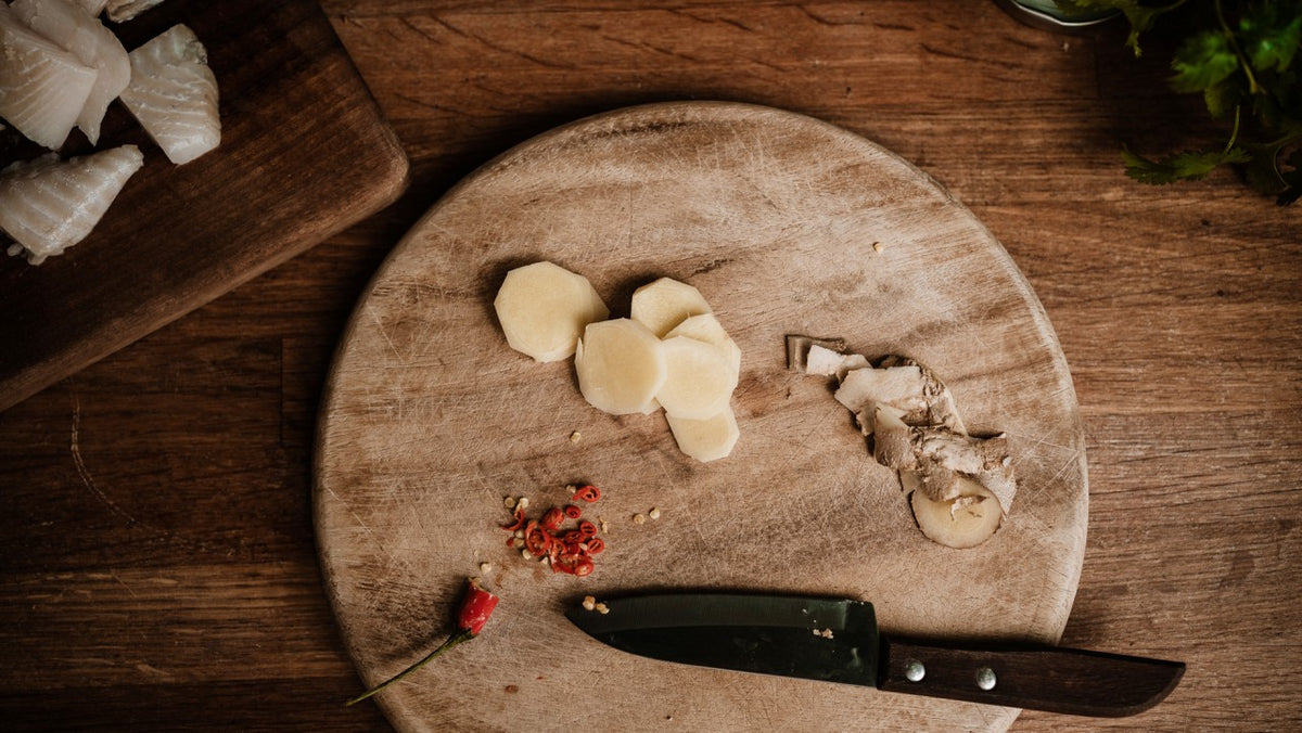 Rundes Schneidebrett mit geschnittenem Ingwer, Chili und Messer auf einem rustikalen Holztisch.