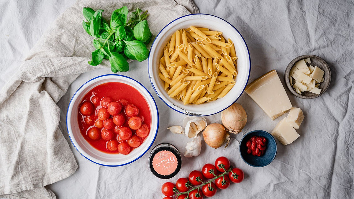 Penne, Tomaten, Käse, Zwiebeln, Basilikum und Knoblauch auf einem weißen Tuch angerichtet.