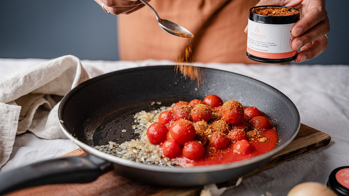 Tomaten und Zwiebeln braten in einer Pfanne auf einem Holzbrett, während eine Person Gewürze darüber streut.