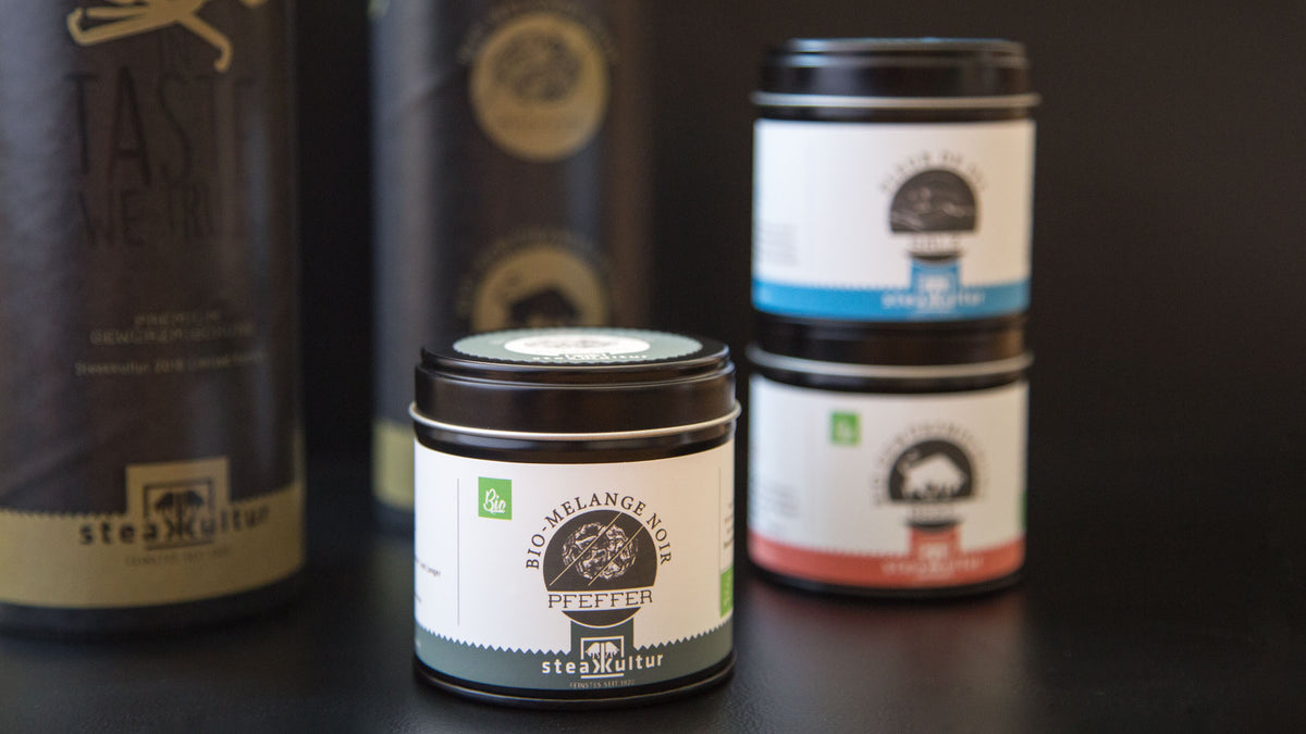 Drei Gewürzgläser mit schwarzen Deckeln, darunter Bio-Melange-Noir-Pfeffer, auf dunklem Untergrund.