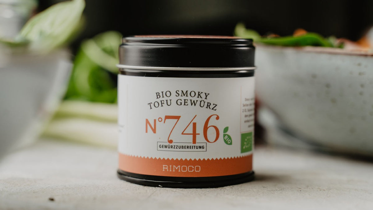 Eine Dose „Bio Smoky Tofu Nr. 746“ steht auf einem Tisch, im Hintergrund sind verschwommene grüne Pflanzen zu sehen.