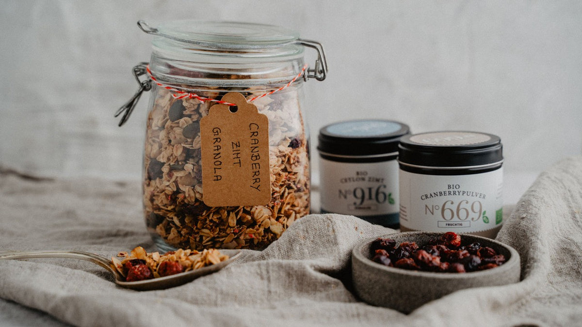 Cranberry-Zimt-Müsli im Glas, dazu Schalen voller Müsli und Cranberries auf einem Tuch.
