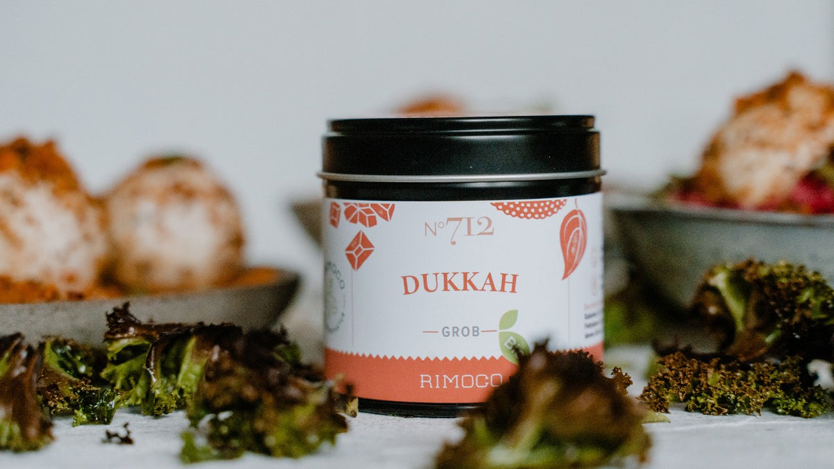 Auf einem Tisch steht eine Dose mit Dukkah-Gewürz neben Blattsalat und frischen Salaten.