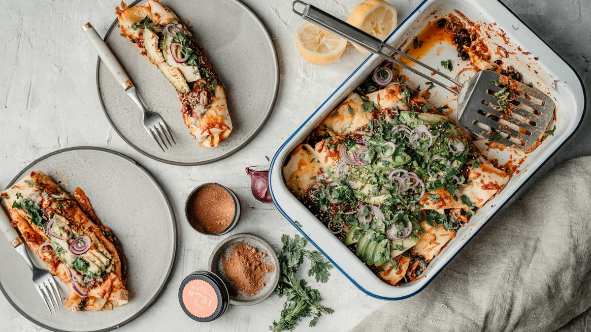 Gebackene Enchiladas mit Avocado, Zwiebeln und Kräutern auf Tellern, dazu Gabeln und Gewürze.