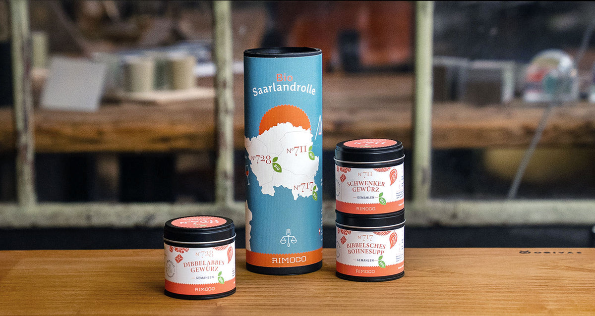 Vier Gewürzgläser und ein hoher zylindrischer Behälter mit der Aufschrift „Bio Saarlandrolle“ auf einem Holztisch.