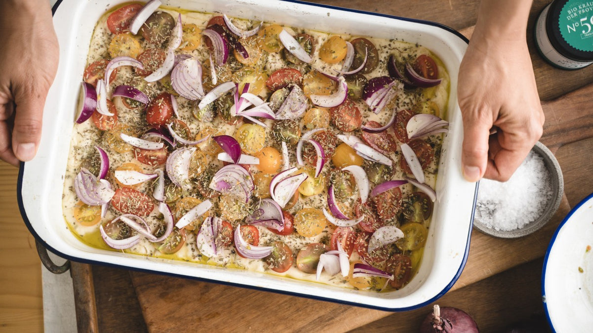 In den Händen wird eine Auflaufform mit Tomatenscheiben, roten Zwiebeln, Kräutern und Olivenöl gehalten.