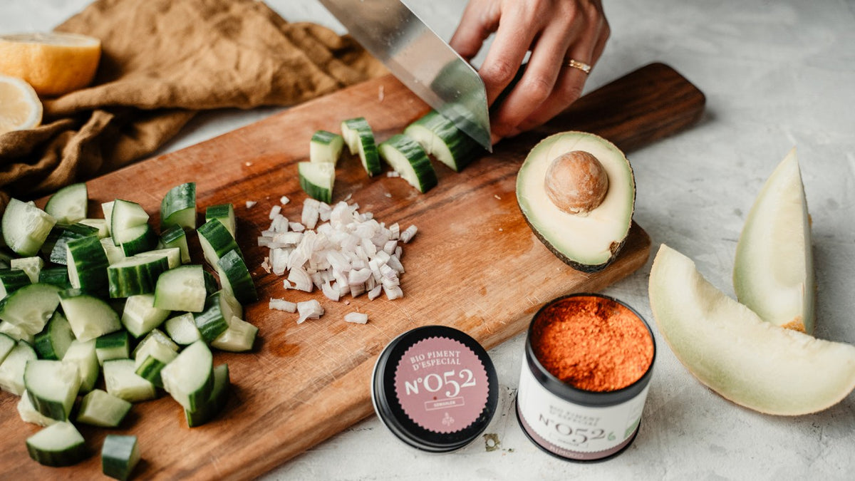 Eine Hand schneidet Gurke und Zwiebel auf einem Holzbrett, daneben liegen Avocado, Melone und gemahlene Gewürze.