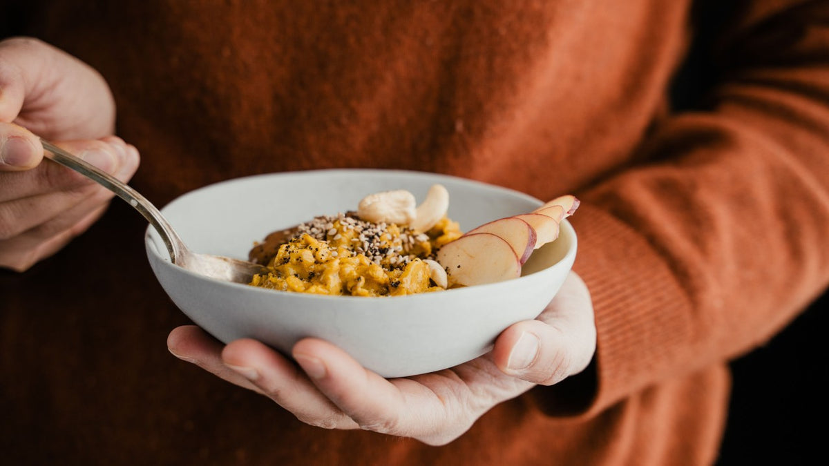 Haferbrei mit Samen, Nüssen und Apfelscheiben in einer Schüssel, die von einer Person im orangefarbenen Pullover gehalten wird.