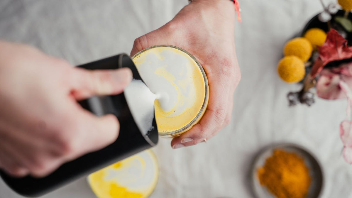 Gelber Becher mit Kurkuma-Latte, ein Barista gießt Milchschaum auf die helle Oberfläche.