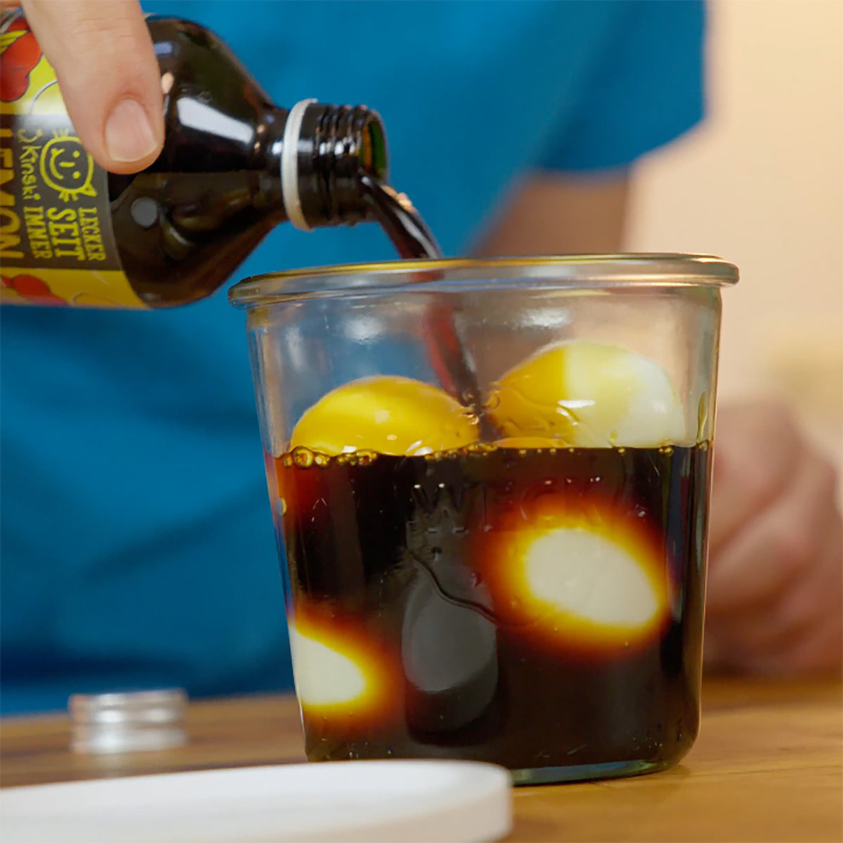 Gekochte Eier in einem Glas, über die Bio Lemon Shoyu Tare gegossen wird, auf Holzoberfläche.