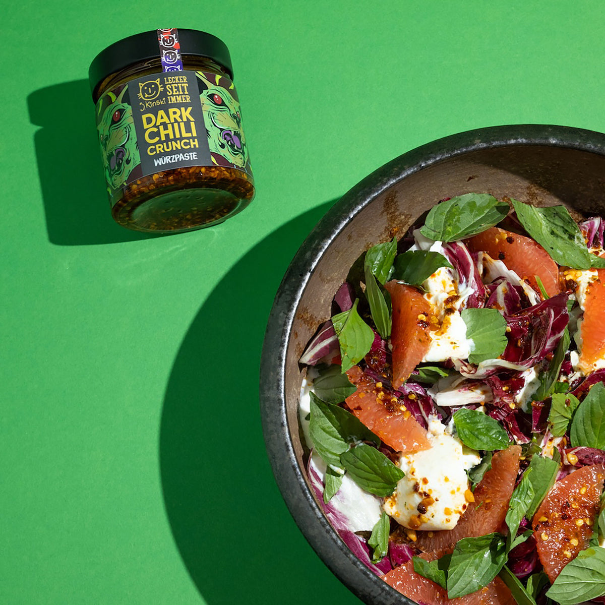 Salat mit Blättern, Zitrusfrüchten, Käse und einem Glas Bio Dark Chili Crunch auf grünem Hintergrund.