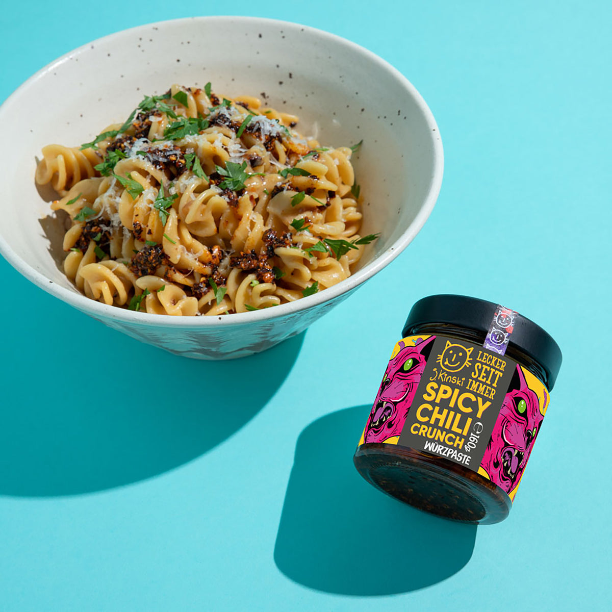 Bio Spicy Chili Crunch im Glas neben Pasta in einer Schale auf blauem Hintergrund.