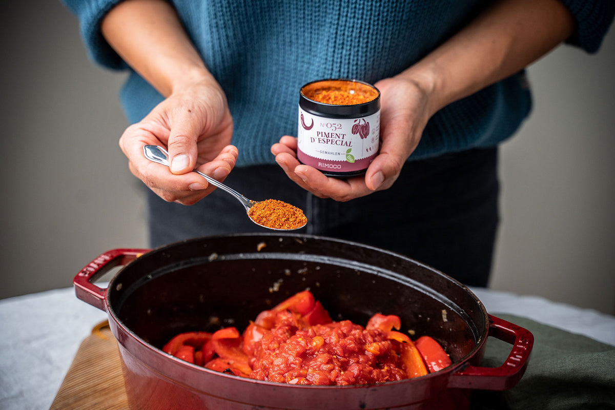 Jemand rührt einen Löffel Gewürzpulver in köchelnde Tomaten und bunte Paprika.