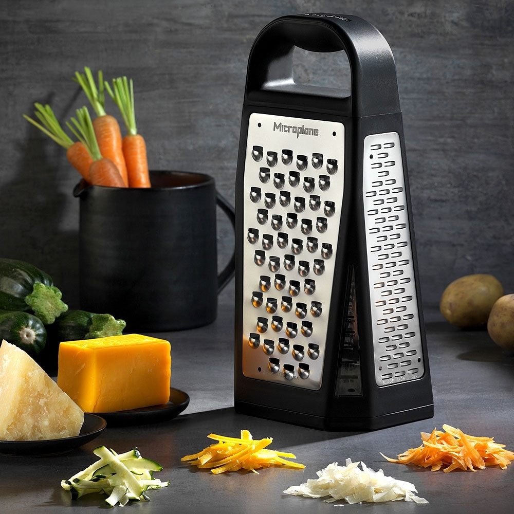 Microplane Turmreibe „Elite Box Grater“, umgeben von geriebenem Gemüse und Käsestückchen.