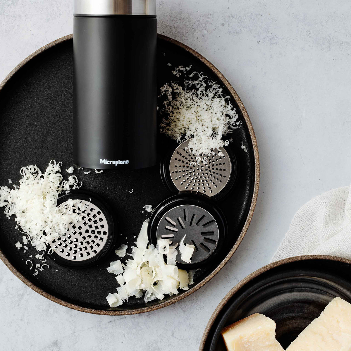 Microplane Universalmühle mit Edelstahlklingen, Aufsätzen, geriebenem Käse und Stücken auf schwarzem Teller.