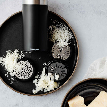 Microplane Universalmühle mit Edelstahlklingen, Aufsätzen, geriebenem Käse und Stücken auf schwarzem Teller.