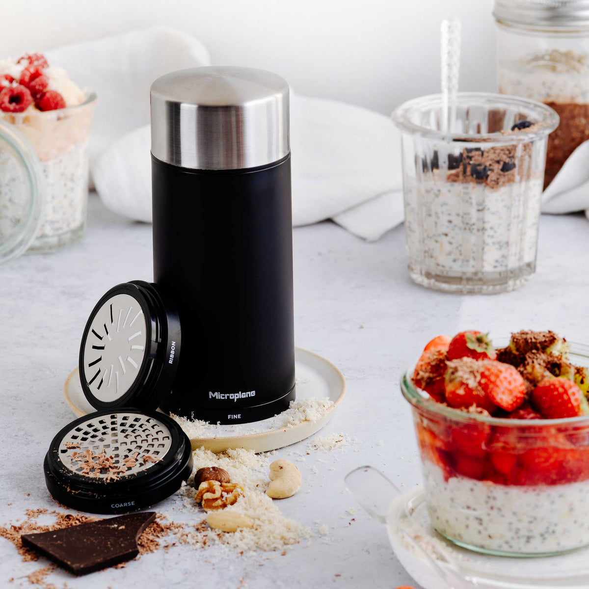 Microplane Universalmühle mit schwarzem Behälter, Edelstahlklingen, Nüssen und geriebenem Essen.