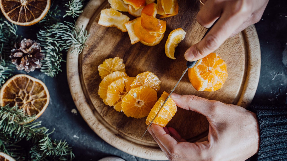 Auf einem Holzbrett schneiden Hände eine frische Orange auf, daneben liegen Schalen und getrocknete Orangenscheiben.