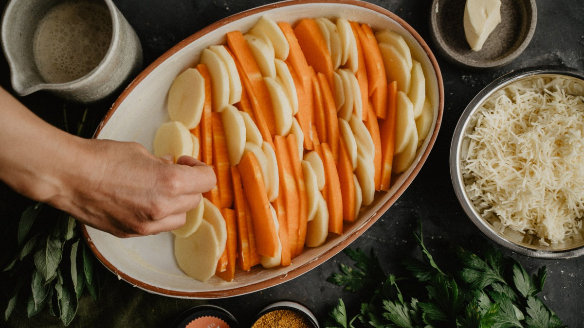 Eine Hand legt Kartoffel- und Karottenscheiben in eine Auflaufform, Käse und Kräuter daneben.