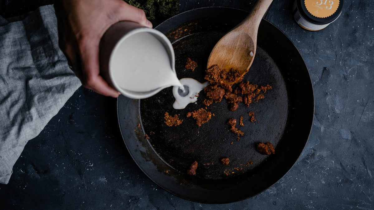 Die Milch wird in eine Pfanne mit den braunen Stücken gegossen und mit einem Holzlöffel umgerührt.