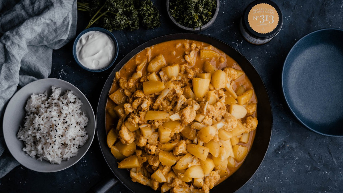 Eine Pfanne mit Kartoffelcurry, dazu weißer Reis und Joghurt auf einem dunklen Holztisch.