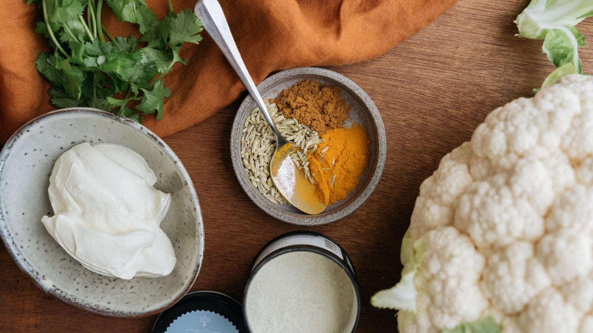 Eine Schale Joghurt, frische Kräuter, gemahlene Gewürze und Blumenkohl auf einem Holzbrett.