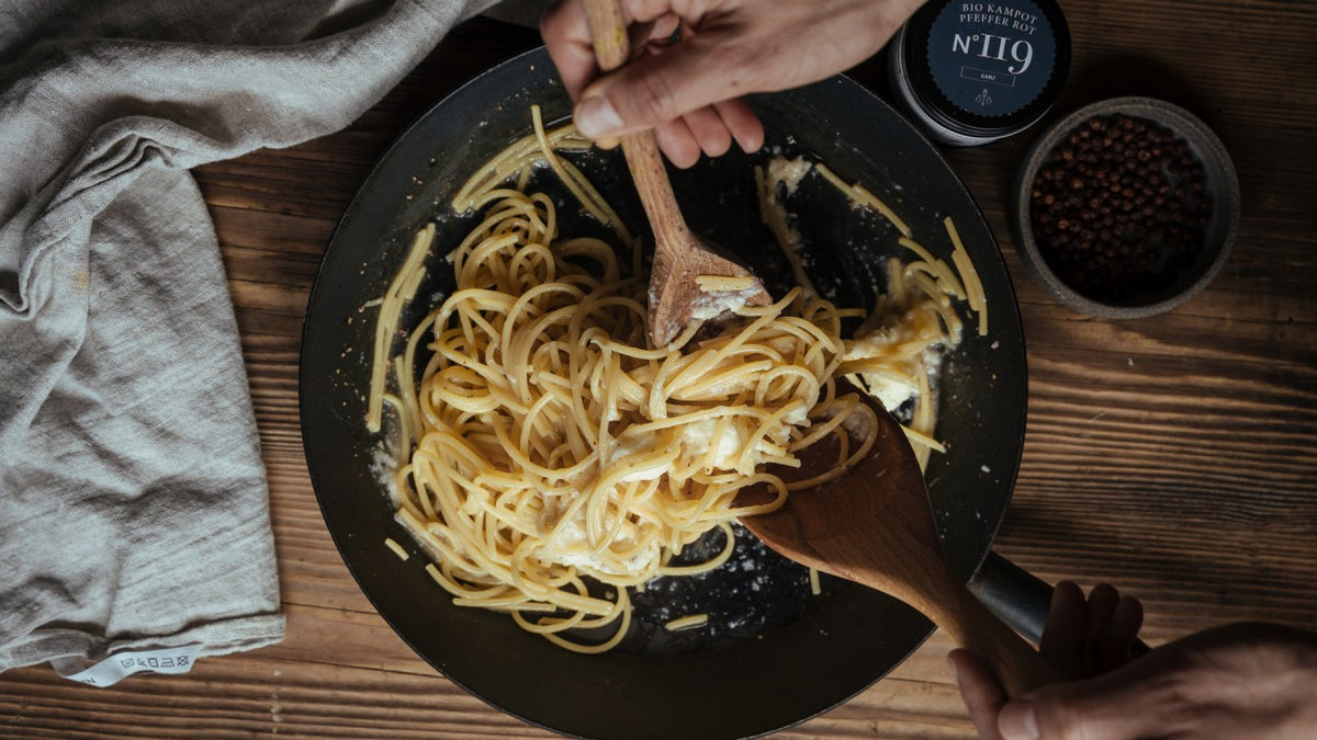 Zwei Hände vermengen Spaghetti mit Käse in einer schwarzen Pfanne auf einem rustikalen Tisch, wobei sie Holzutensilien verwenden.