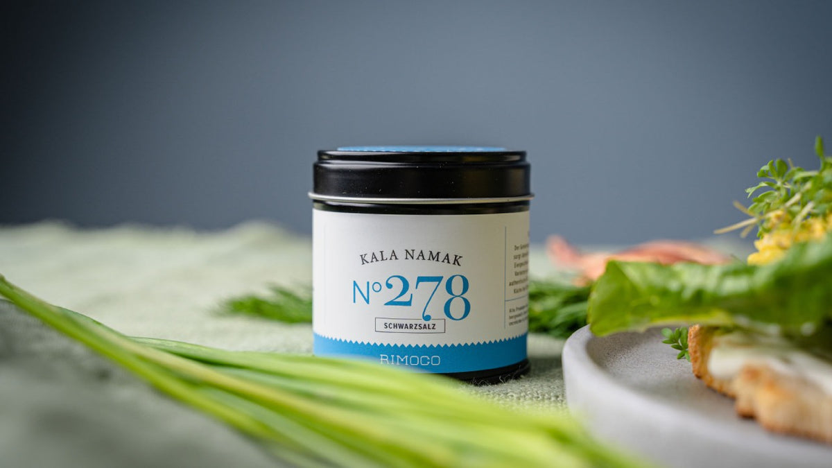 Kala Namak Nr. 278 steht auf einem Tisch neben Blattgemüse und essbaren Blüten.