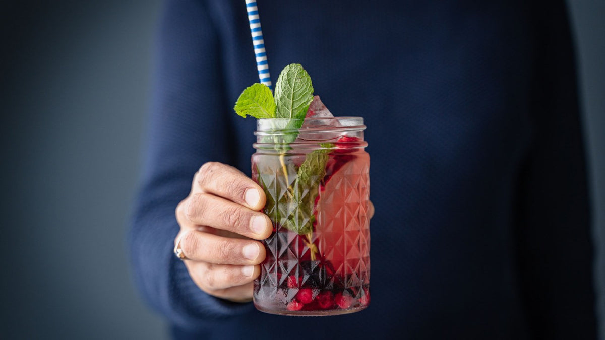 Eine Person hält ein Glas mit Beerensaft, frischer Minze, Eiswürfeln und einem gestreiften Strohhalm in der Hand.