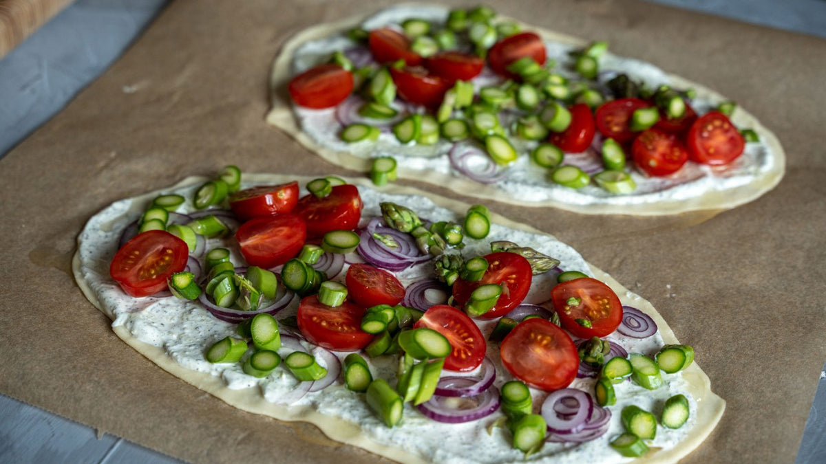 Fladenbrote mit Creme, Kirschtomaten, roten Zwiebeln und Spargel auf Backpapier.