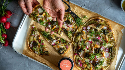 Hände halten ein Stück Gemüsepizza, daneben frische Radieschen und Pesto-Gewürze auf Backpapier.