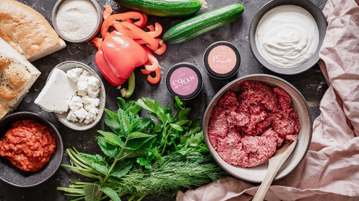 Frische Zutaten: Hackfleisch, Kräuter, Paprika, Gurken, Käse, Saucen und frisches Brot.