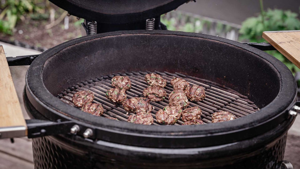 Fleischbällchen brutzeln auf einem Grill in einem runden schwarzen Barbecue-Smoker im Freien.