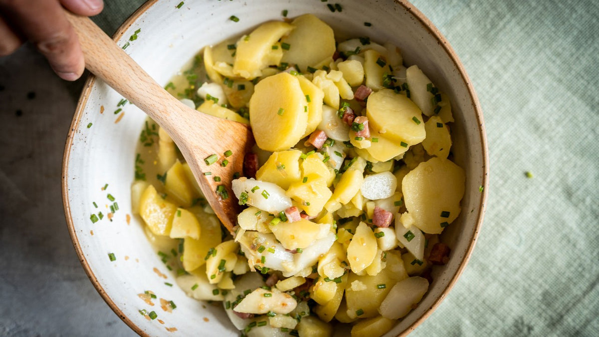 Kartoffelsalat mit Zwiebeln, knusprigem Speck und Schnittlauch in einer Schüssel mit einem Holzlöffel.
