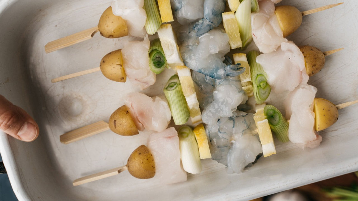Spieße mit Kartoffeln, Garnelen, Fisch und Gemüse liegen in einer weißen Auflaufform.