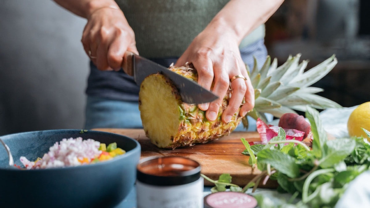 Eine Person schneidet eine reife Ananas auf einem Holzbrett auf, umgeben von frischen tropischen Zutaten.