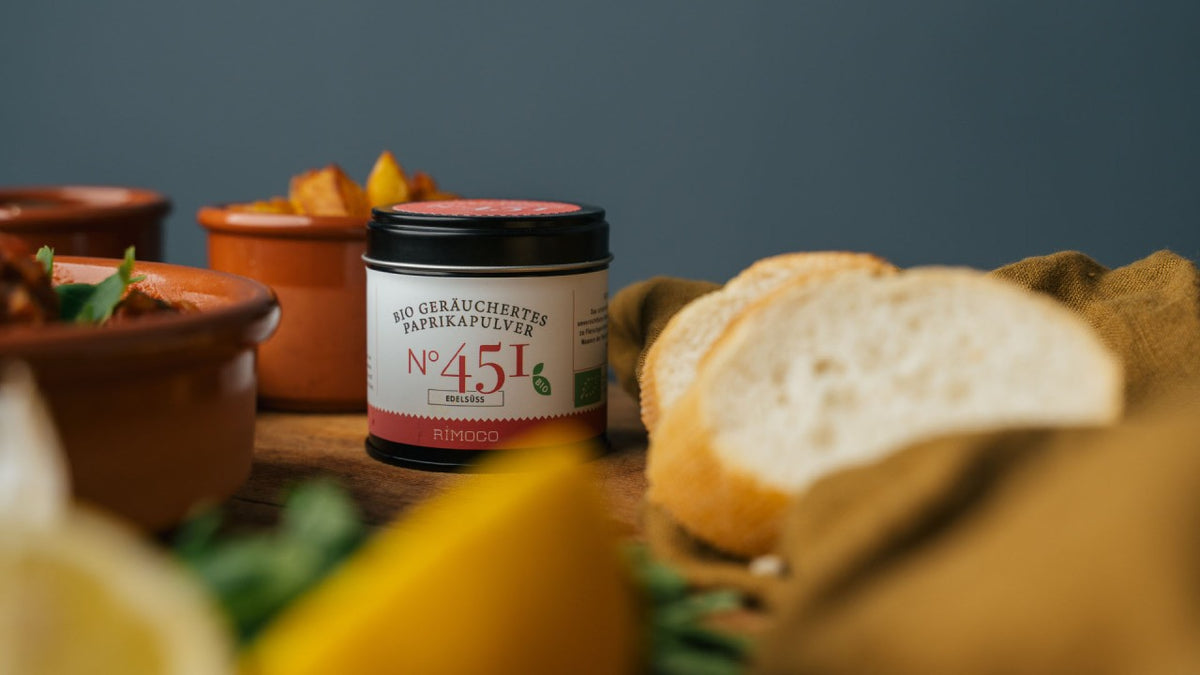 Paprikapulver im Glas neben Brotscheiben und Tellern auf einem rustikalen Holztisch.