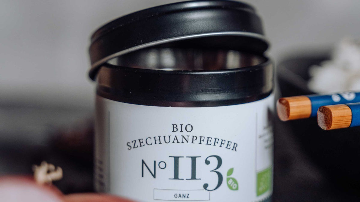 Schwarzes Glas mit der Aufschrift „Bio Szechuanpfeffer No II3“, Deckel teilweise geöffnet, Inhalt sichtbar.