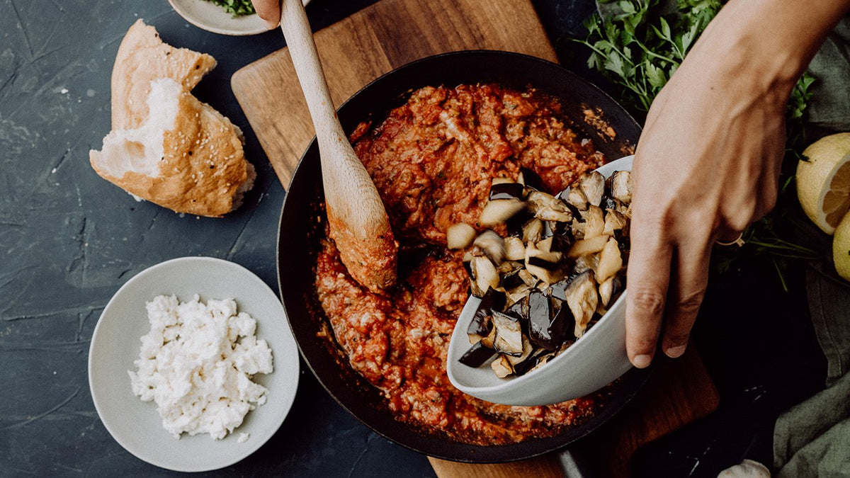 Die in Scheiben geschnittene Aubergine wird in eine Pfanne mit Tomatensoße gegeben; Brot und Käse stehen auf dem Tisch.