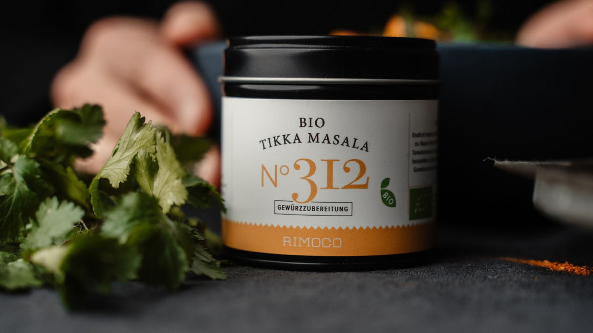 Bio-Tikka-Masala-Gewürzmischung im Glas, frischer Koriander und eine unscharfe Hand im Hintergrund.