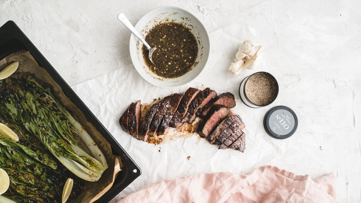Gegrilltes Steak mit grobem Meersalz, Sauce und gegrilltem Gemüse auf weißer Oberfläche.