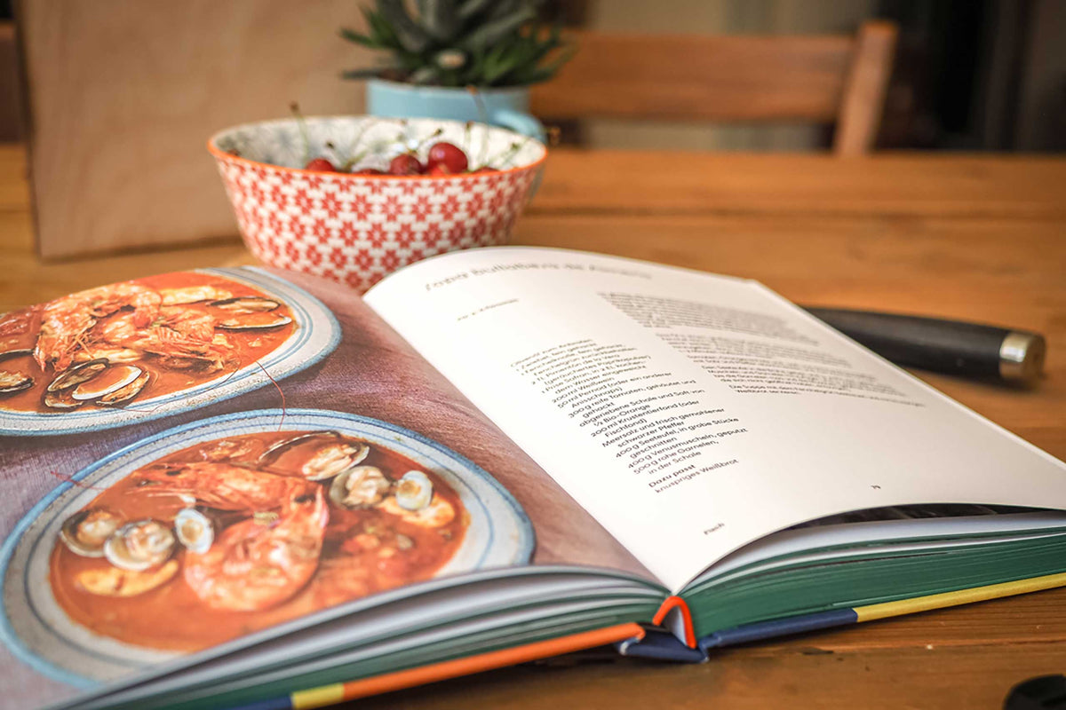 Aufgeschlagenes Kochbuch mit Meeresfrucht-Rezept, daneben eine Schale Kirschen mit Muster.