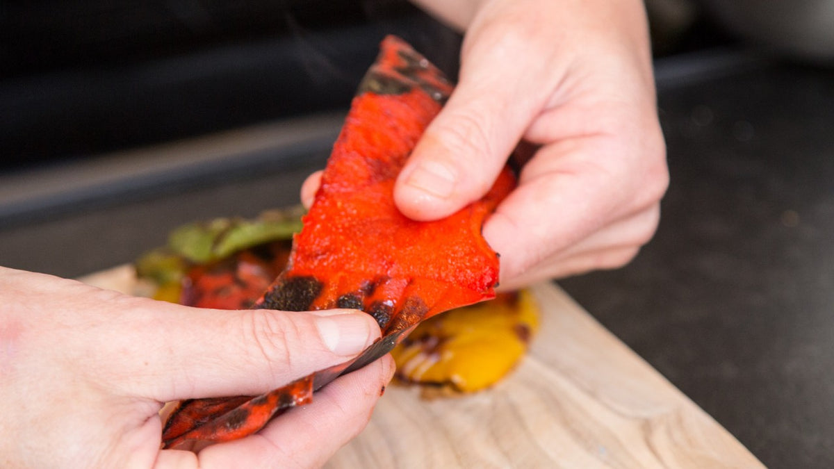 Hände ziehen die verkohlte Haut von einer gerösteten roten Paprika auf einem Holzbrett ab.
