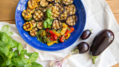 Gegrilltes Gemüse mit Basilikum, Aubergine und Knoblauch in blauer Schale, dazu Balsamico-Flasche.