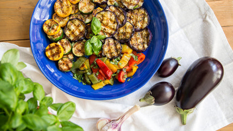 Gegrilltes Gemüse mit Basilikum, Aubergine und Knoblauch in blauer Schale, dazu Balsamico-Flasche.