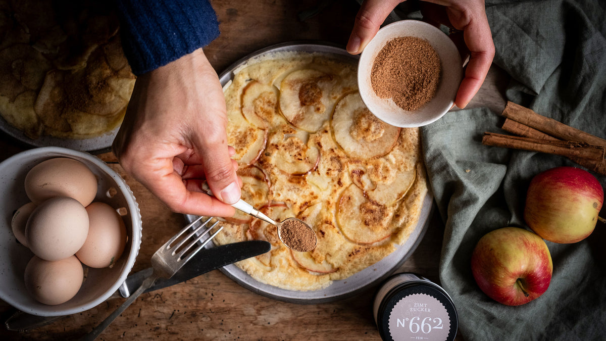 Apfelpfannkuchen, bestreut mit gemahlenem Zimt, umgeben von frischen Äpfeln und Zimtstangen.