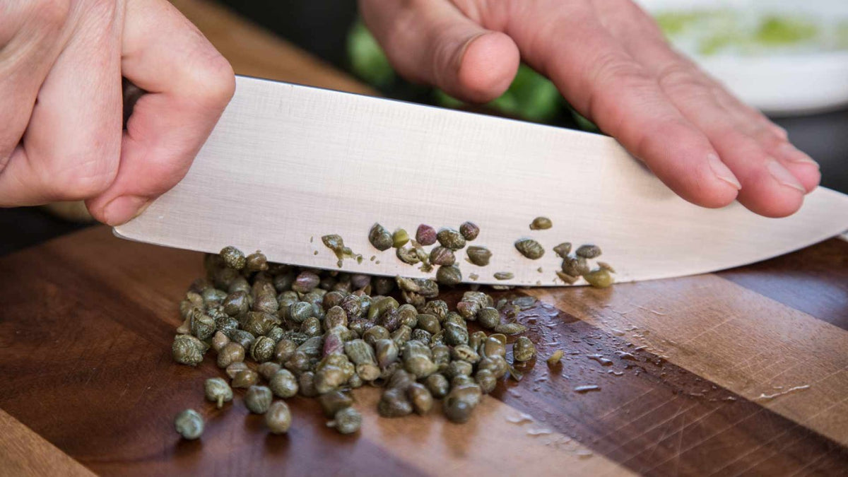 Eine Hand hackt Kapern mit einem scharfen Messer auf einem Holzschneidebrett fein.