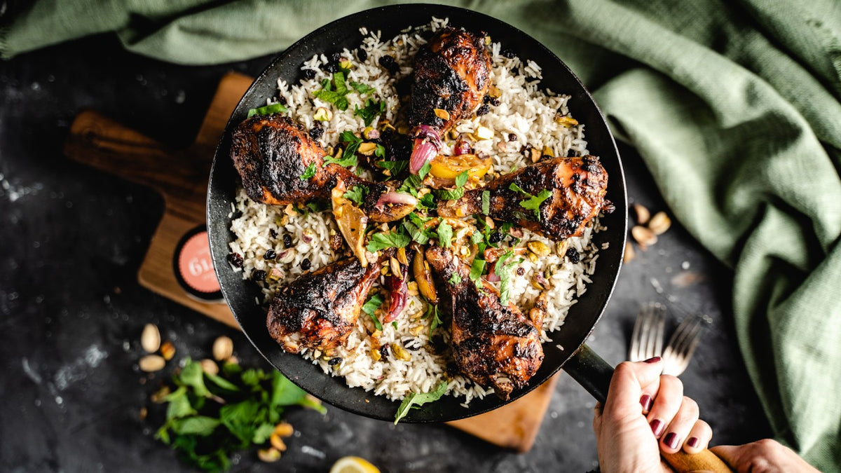 Eine Hand hält eine Pfanne mit gegrillten Hähnchenschenkeln, Reis, Kräutern und buntem Gemüse.