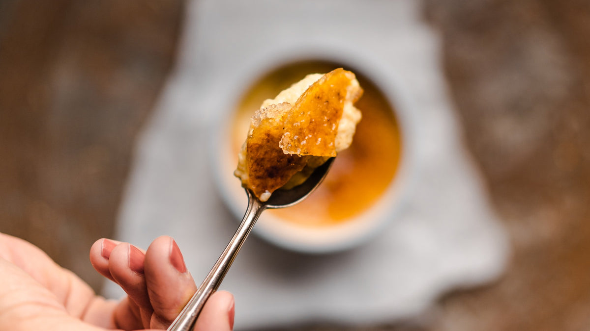 Eine Hand hält einen Löffel mit cremiger Crème brûlée und karamellisierter Zuckerkruste über die Schale.